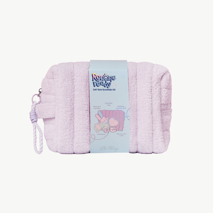 A light lilac plush cosmetic pouch with a matching wrist strap, wrapped in a light blue paper band featuring the 'Routinely Ready' brand logo and product details.