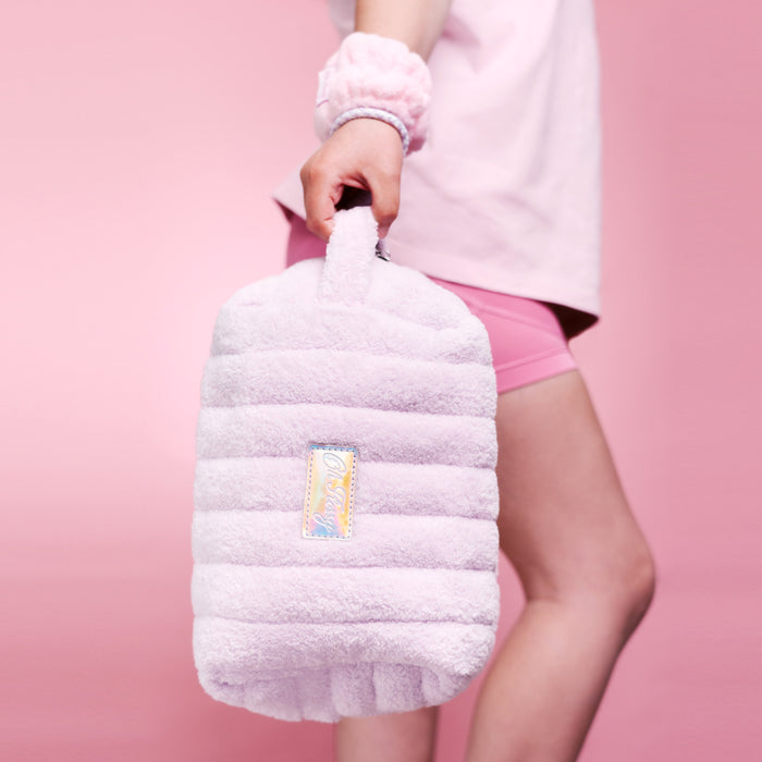 A person in a pink outfit holds a light lilac ribbed plush cosmetic bag with a small iridescent logo patch against a solid pink background.